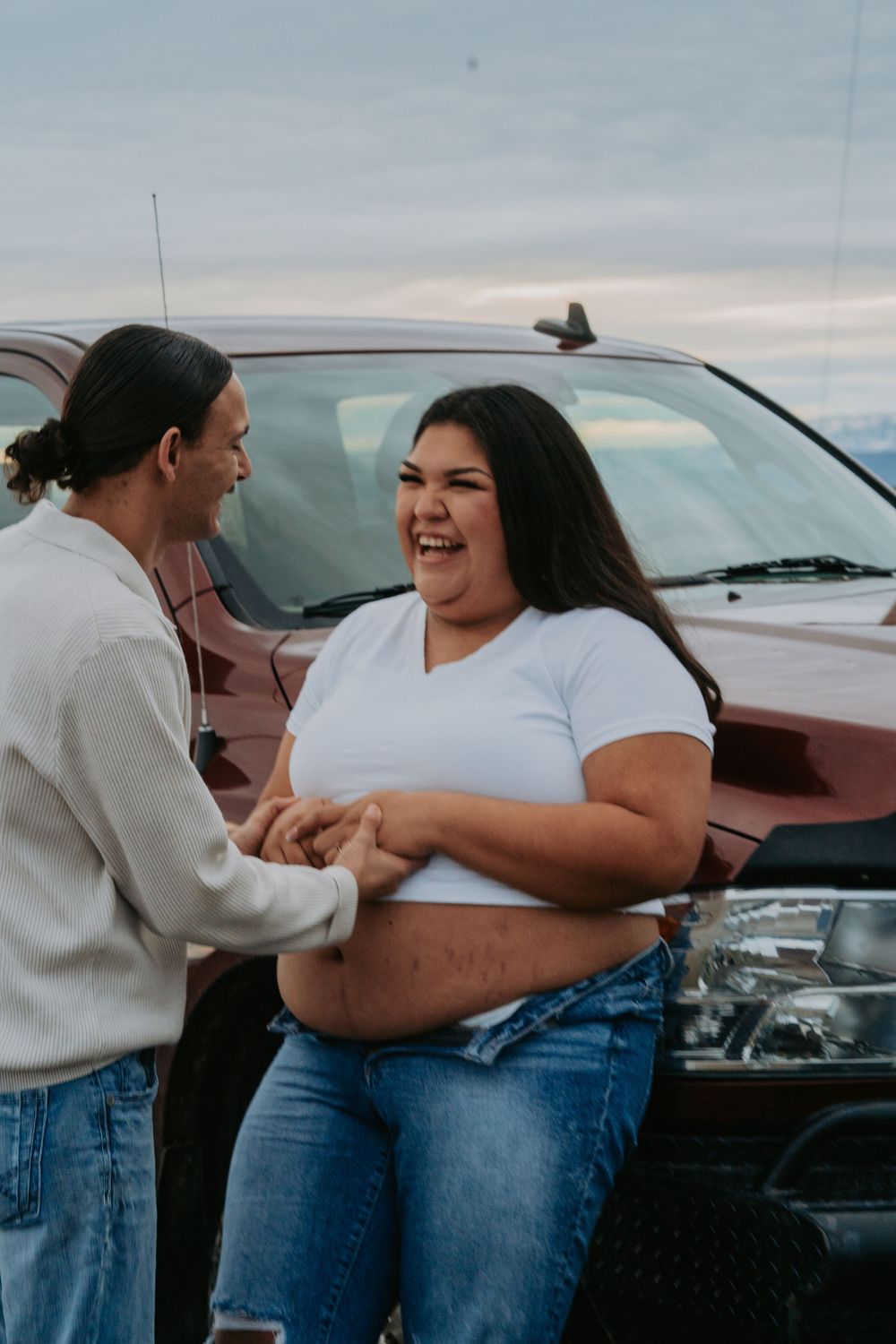 A laughing couple holds hands in front of a red truck outdoors, woman in white crop top and jeans.