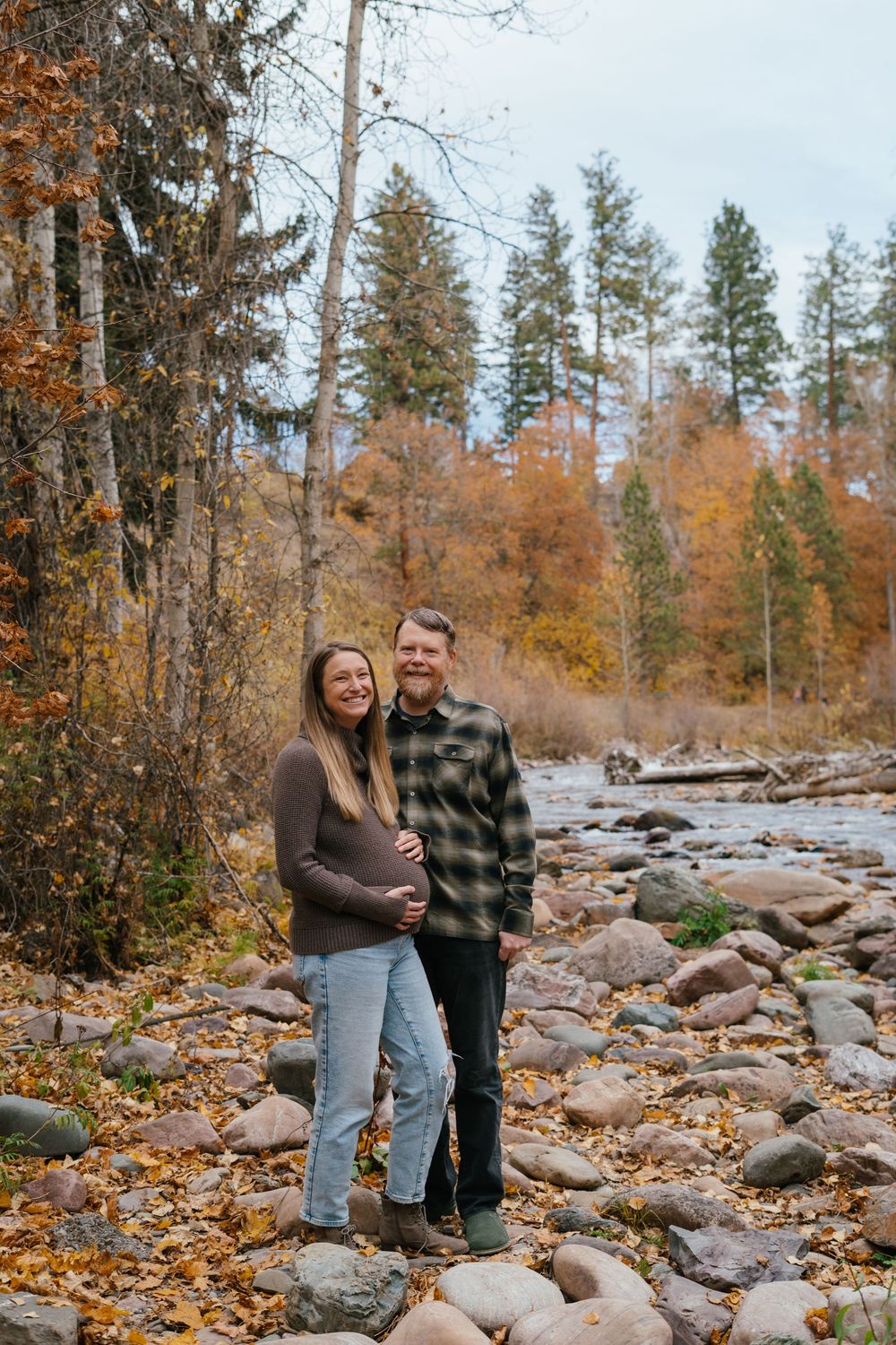 Two people stand together on rocky terrain surrounded by autumn trees and fallen leaves in a natural outdoor setting.