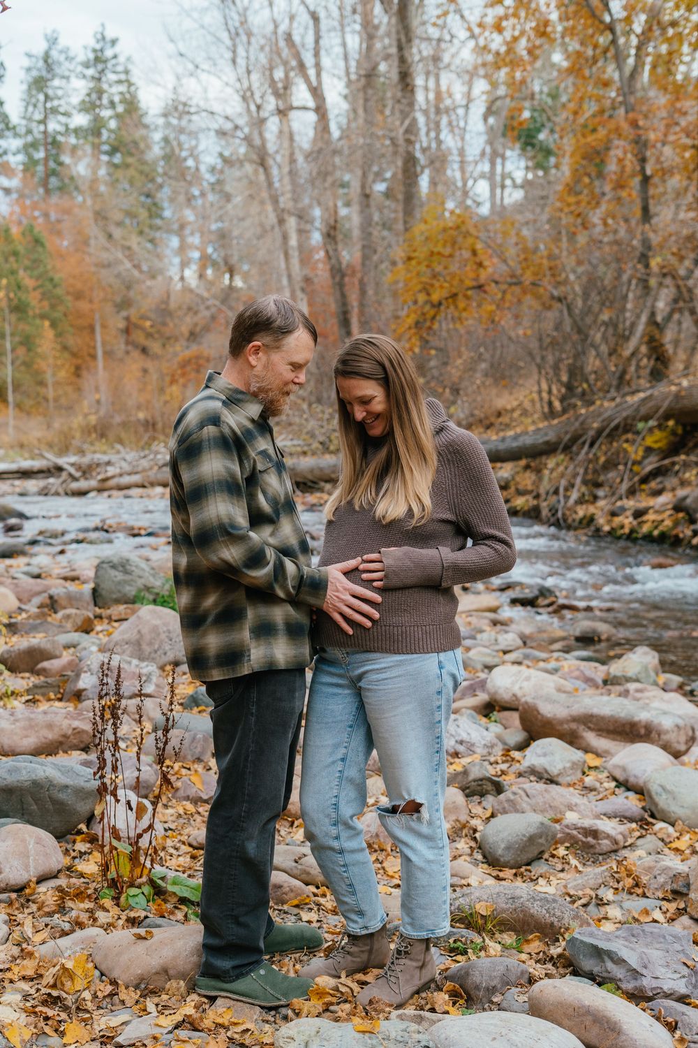 A couple shares an intimate moment by a rocky stream surrounded by autumn foliage and fallen leaves.