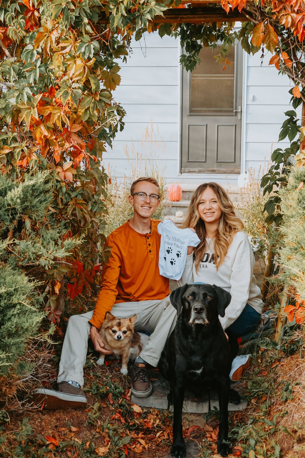 A couple poses on their porch with two dogs surrounded by fall foliage.