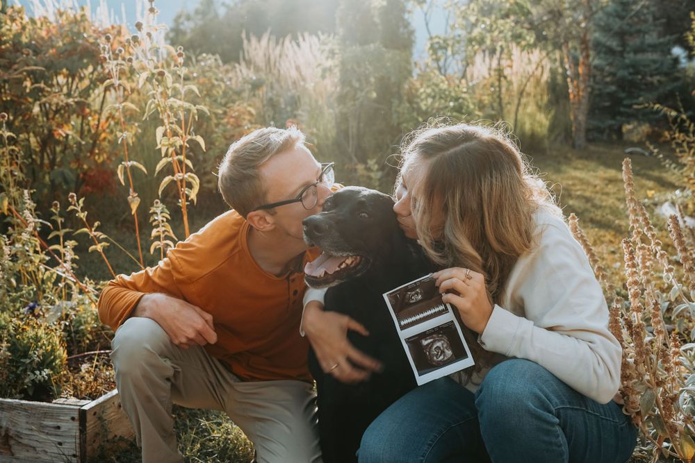 A couple laughs with their small dog while holding a sonogram photo outdoors.