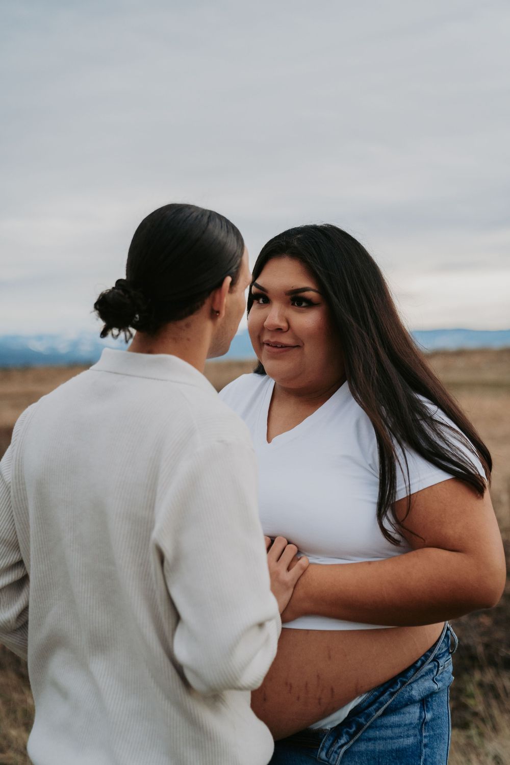 Pregnant woman in white crop top and jeans holds her baby bump while partner faces her in an outdoor field setting.