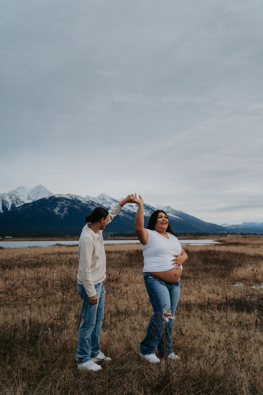 Pregnant couple dancing in a mountain field, woman in white top touching belly, snowy peaks and lake in background.