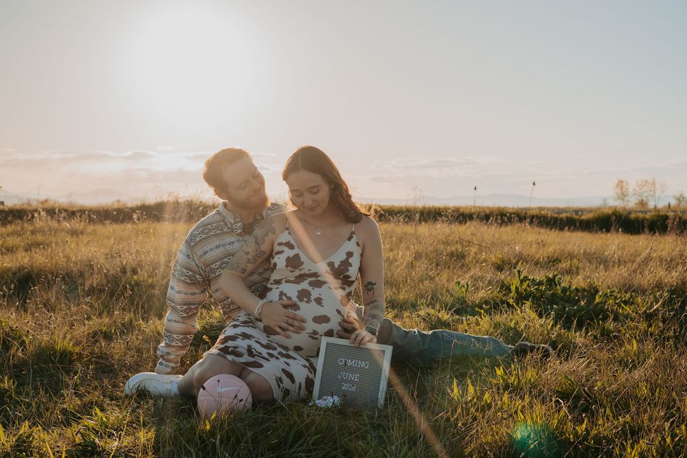 Couple sitting in sunny golden field, woman in floral dress with baby bump, holding a sign during maternity photo session.