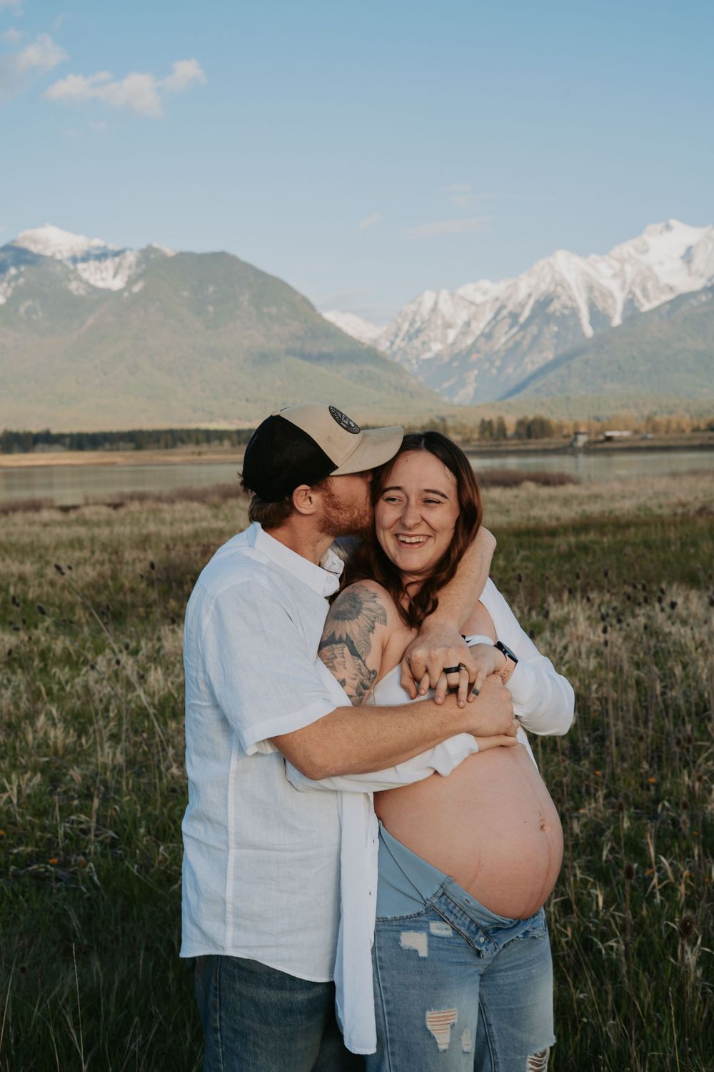 A couple shares a tender moment in a mountain meadow, man kissing pregnant woman's cheek while cradling her baby bump.