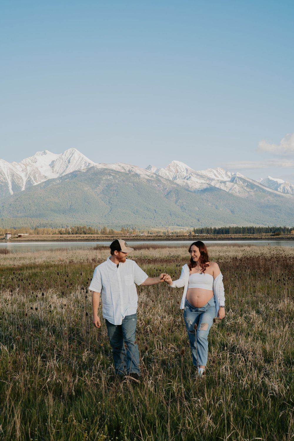 Pregnant couple holding hands walking in a field with snow-capped mountains in the background.