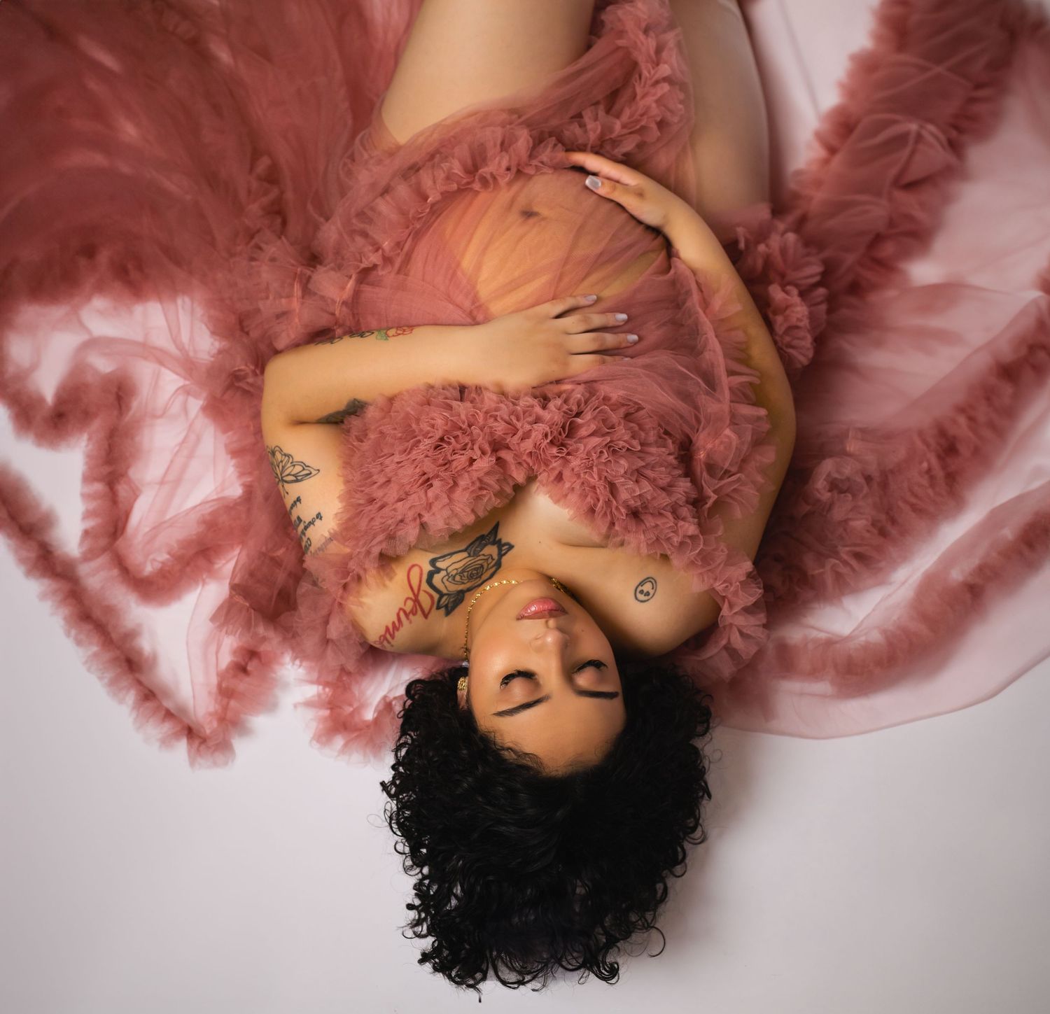 Pregnant woman in pink tulle gown lying down with tattoos visible in artistic maternity portrait.