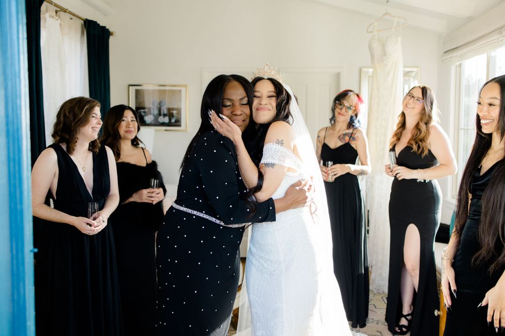 Wedding party members in black dresses and white gown gathered in a bright room.