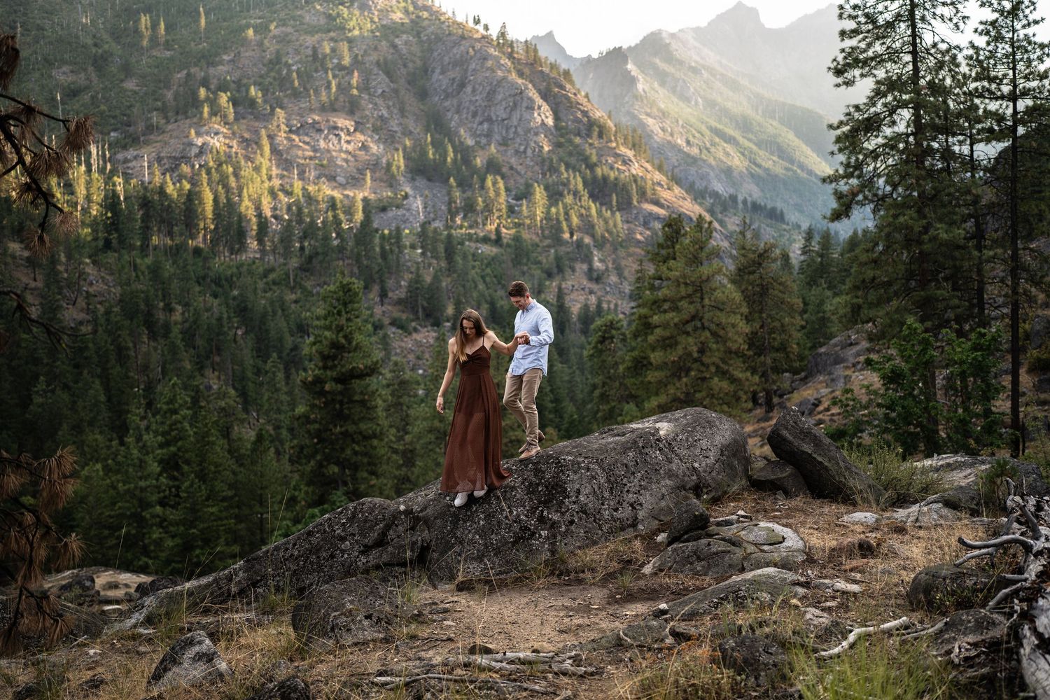 A couple stands on a rocky mountain overlook surrounded by pine trees and distant peaks.