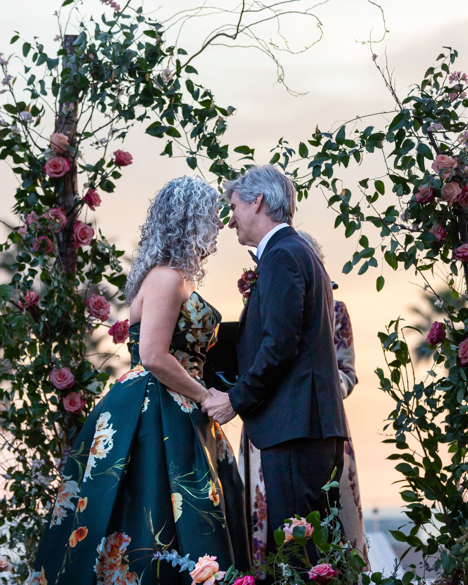 A couple exchanges vows during an outdoor garden wedding ceremony surrounded by pink flowers and greenery at sunset.