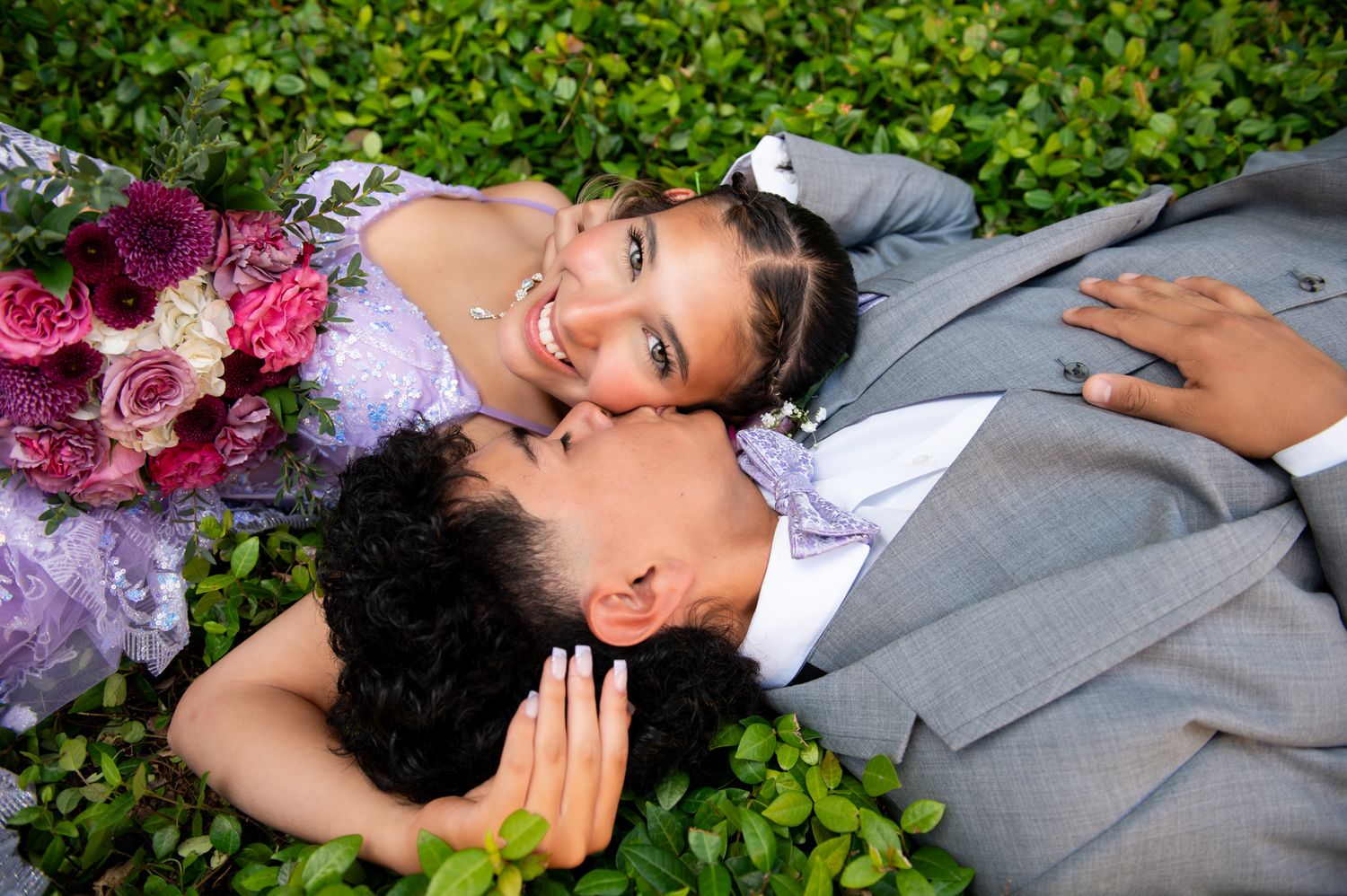 Newlywed couple lying in grass with vibrant pink and purple bouquet at outdoor wedding photoshoot.