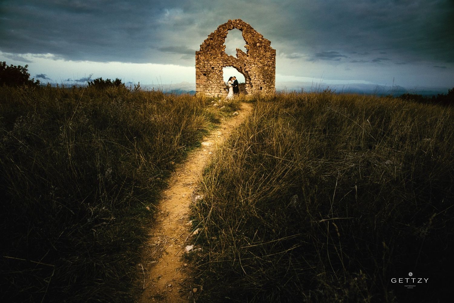 A large rustic wagon wheel stands against a cloudy blue sky while someone in a flowing white dress poses beside it.