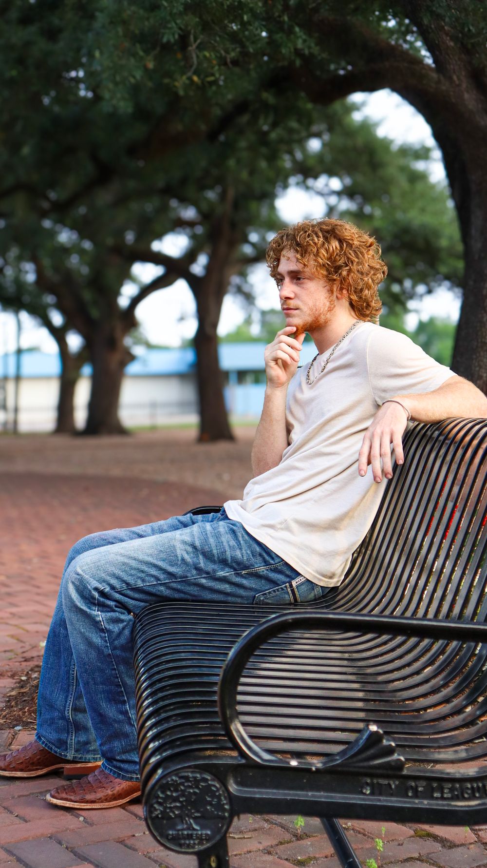 Person sitting on a black metal park bench in a casual pose with trees in the background.