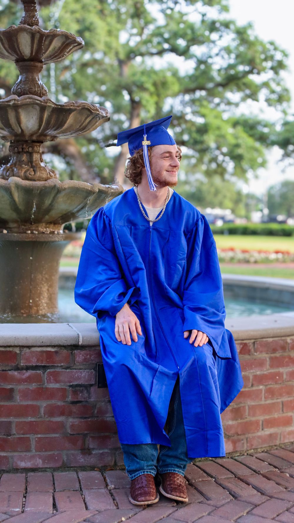 Graduate in blue cap and gown sitting on brick wall during outdoor photo session.