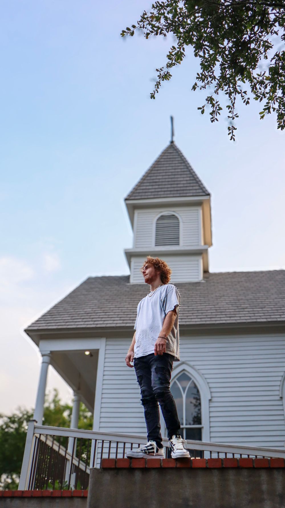 Figure standing on brick ledge in front of white church steeple against blue sky.