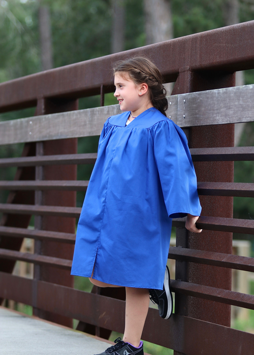 Graduate wearing a bright blue graduation gown walks across a wooden bridge on a sunny day.