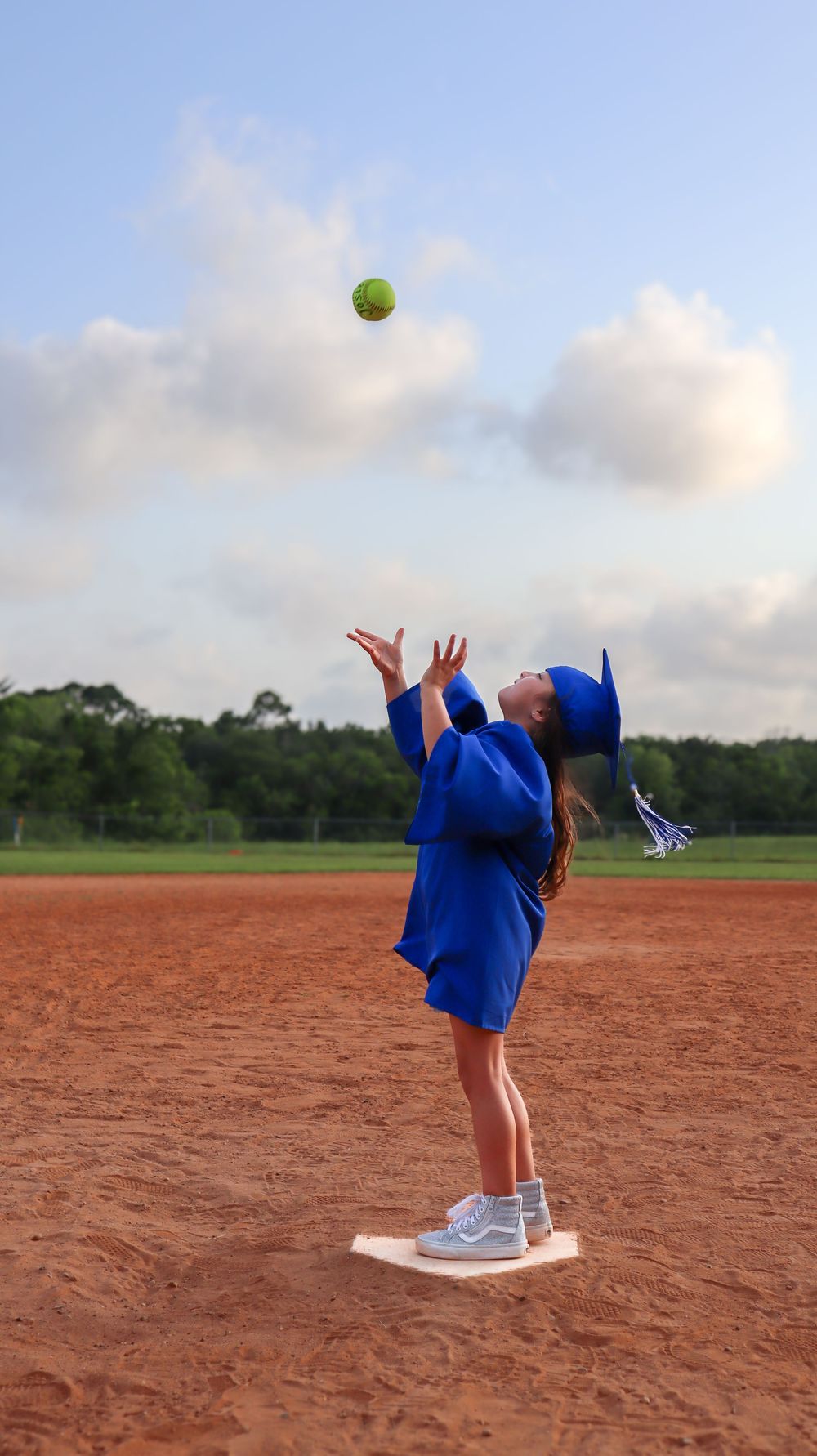 Person in blue graduation gown tosses softball on dirt field.