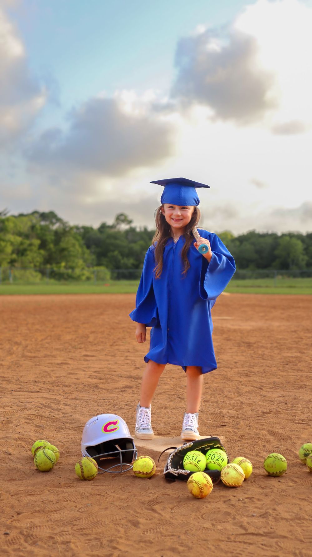 Graduate in blue regalia poses with softballs arranged on dirt field.