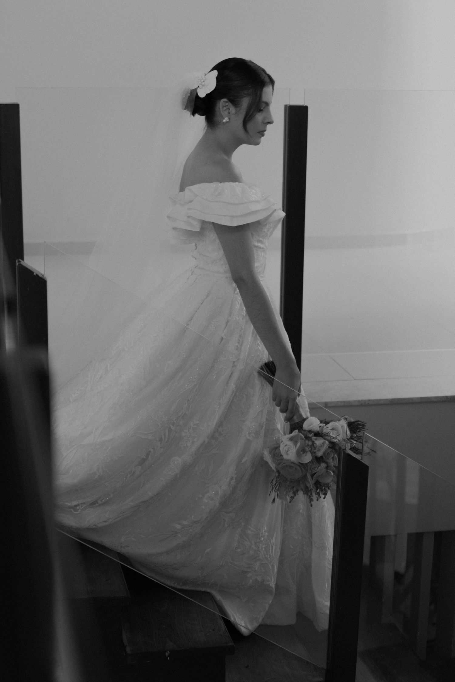 Black and white photo of a bride in an off-shoulder gown standing on stairs.