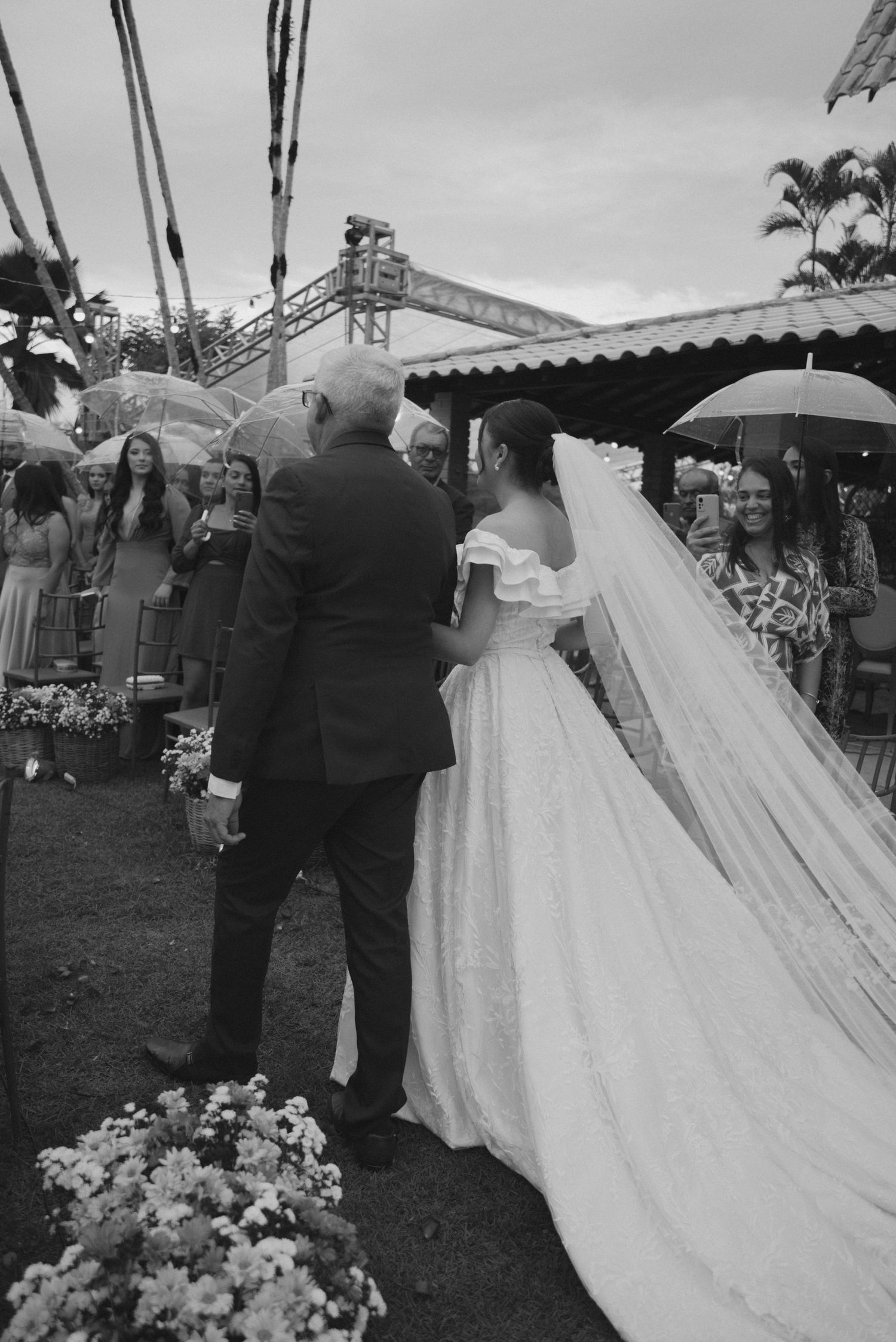 Black and white photo of bride in flowing dress walking down outdoor aisle with umbrellas.