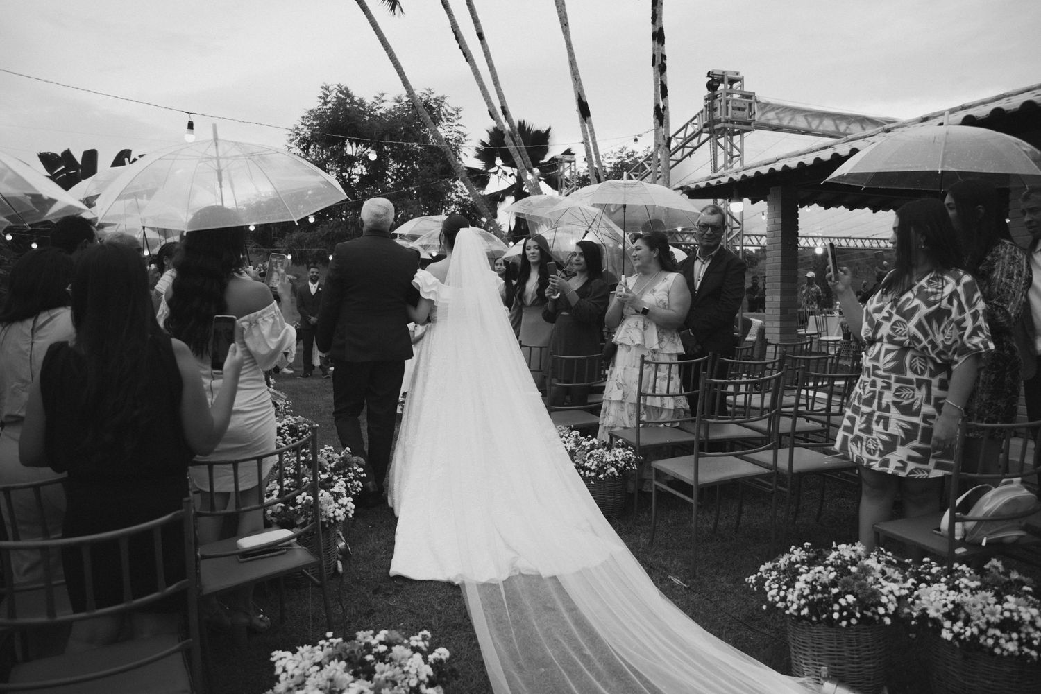 Wedding ceremony under palm trees with guests holding umbrellas in black and white.