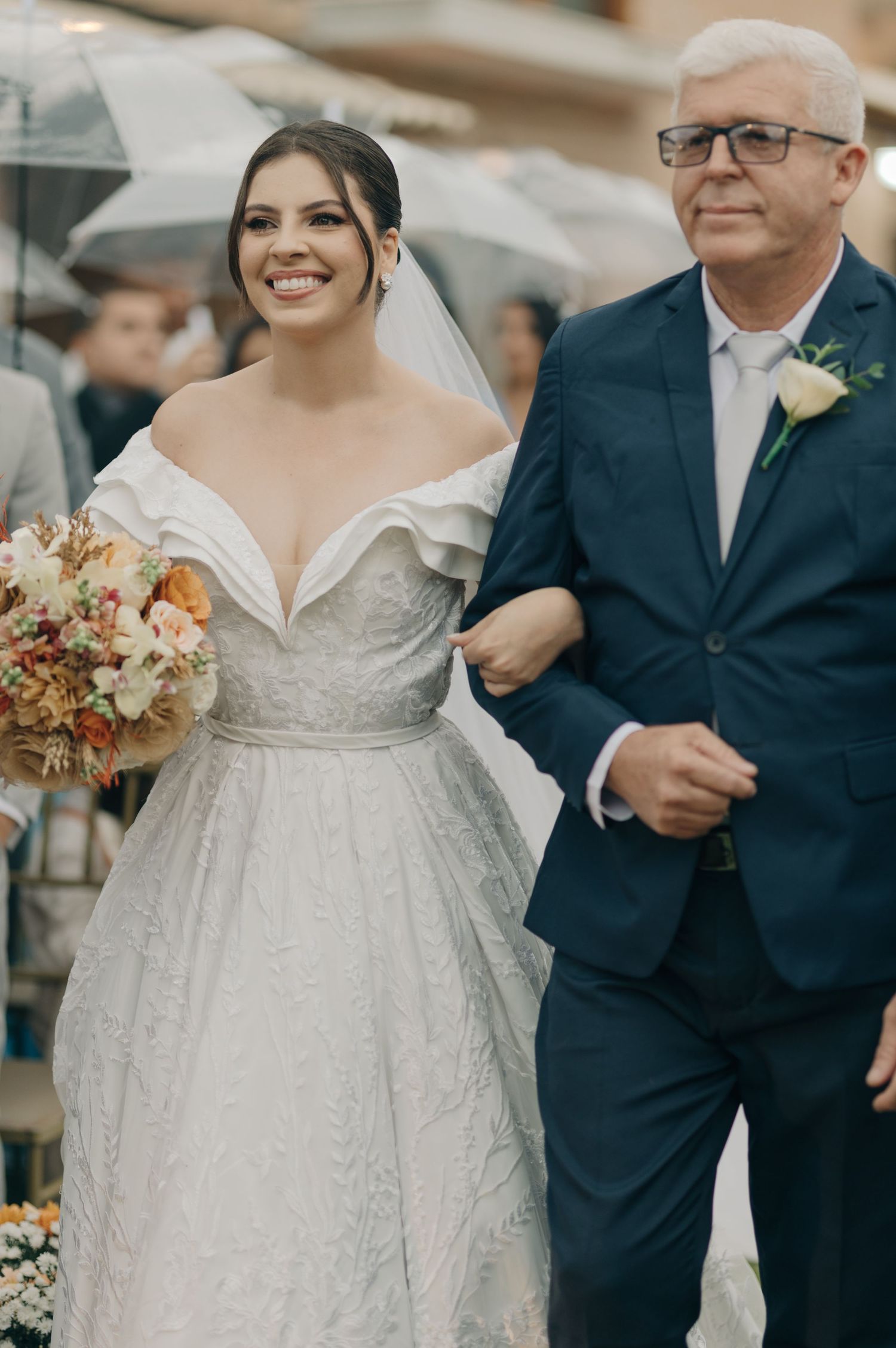 Bride in off-shoulder wedding gown walking with father down outdoor aisle.