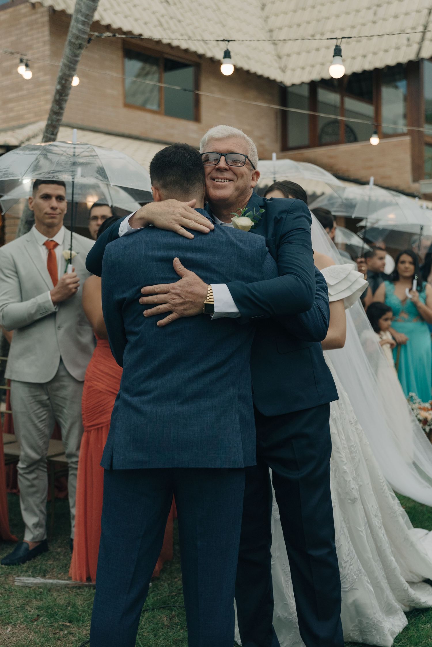 Emotional embrace between two men in suits during outdoor wedding celebration.