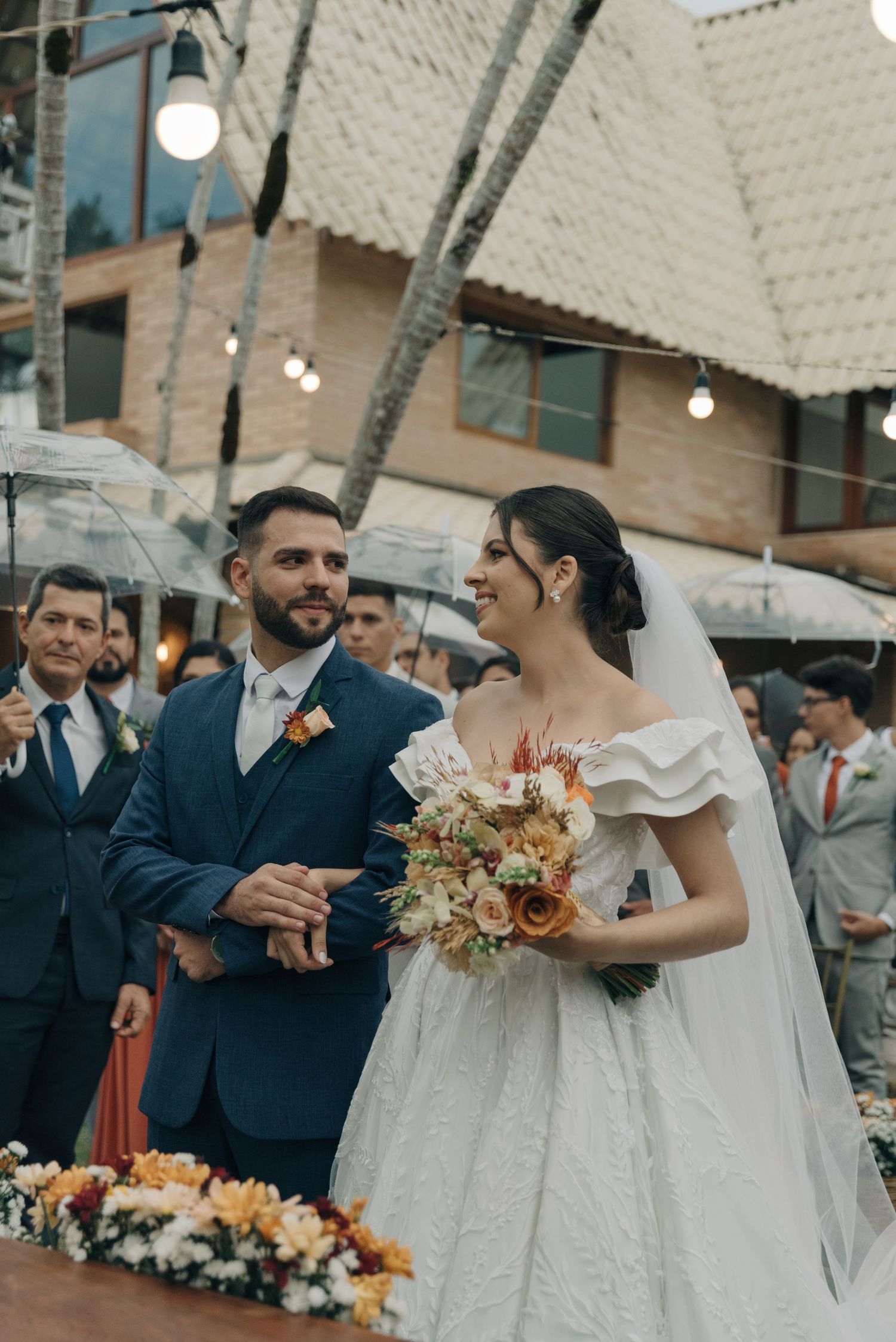 Bride in white gown holding autumn bouquet next to groom in navy suit at outdoor ceremony.