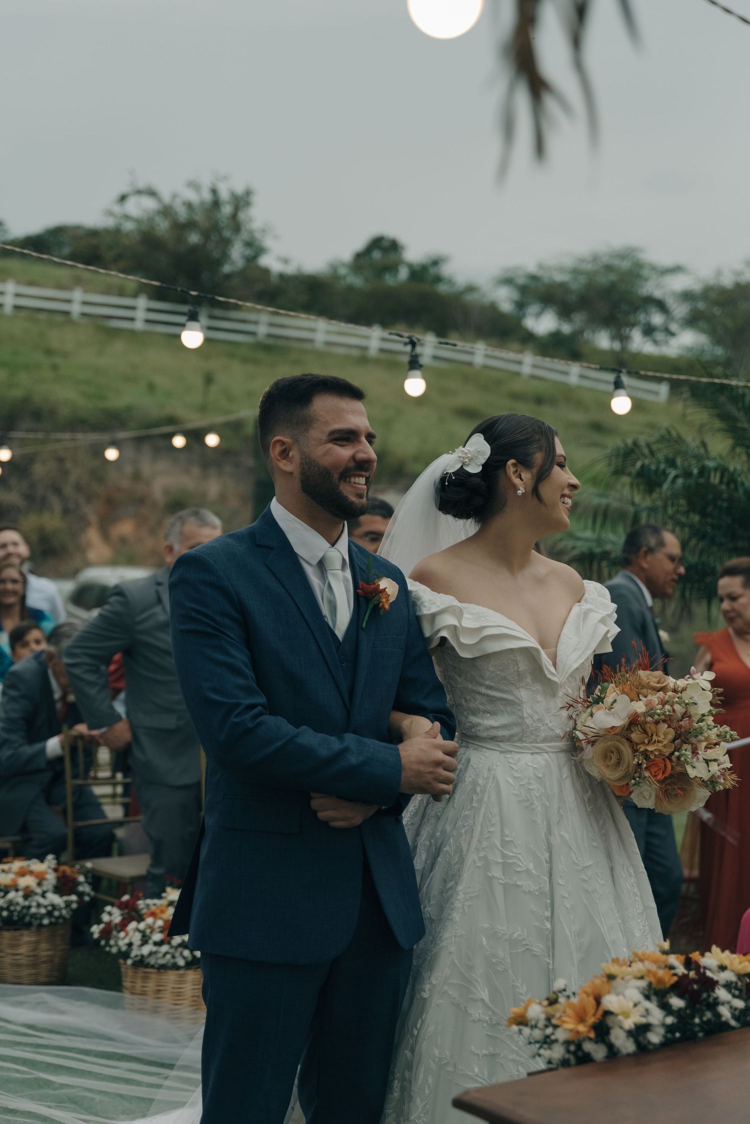 An outdoor wedding celebration on a hilltop with string lights and white fencing in the background.
