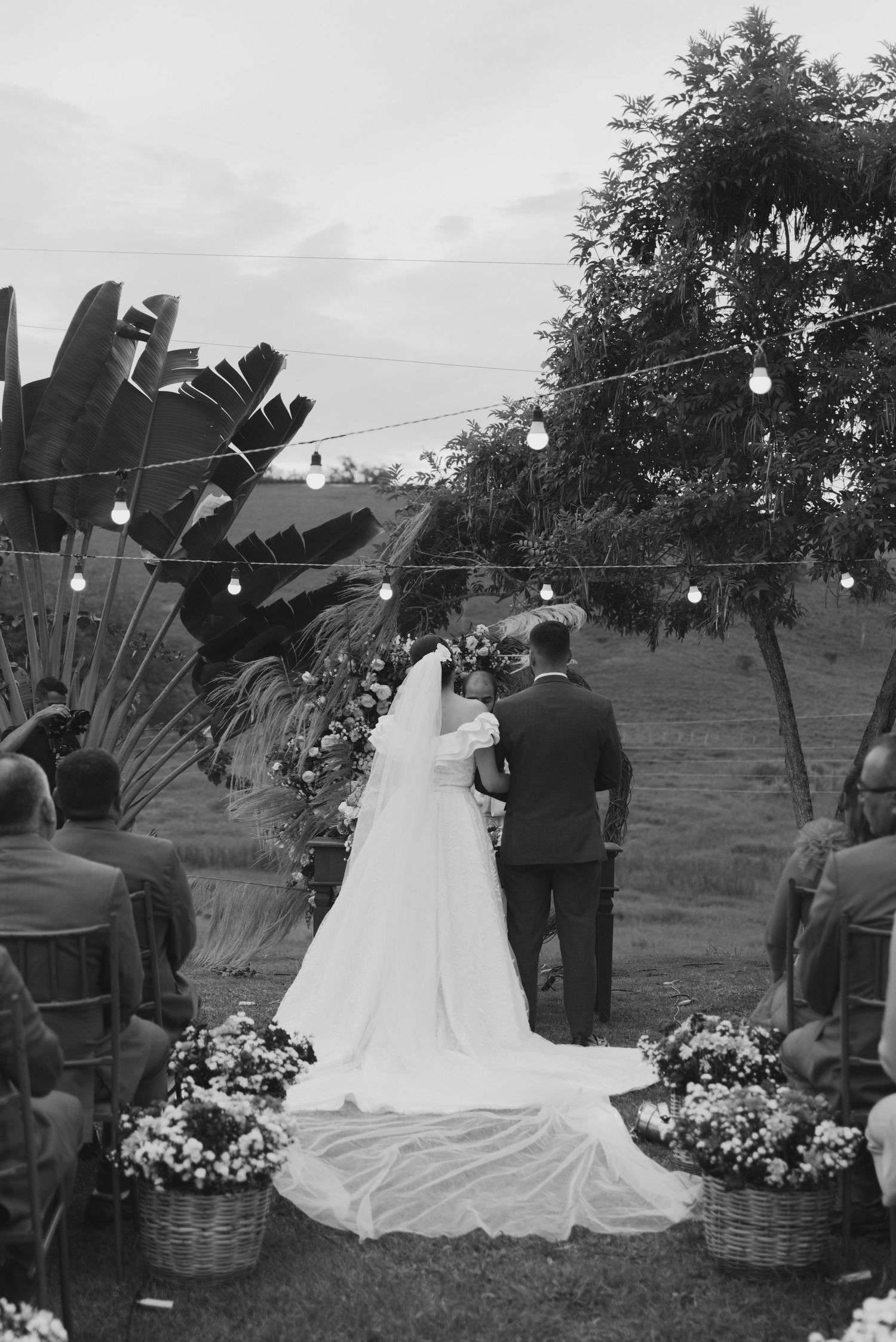 A black and white wedding ceremony photo showing a long train dress and floral aisle decorations.