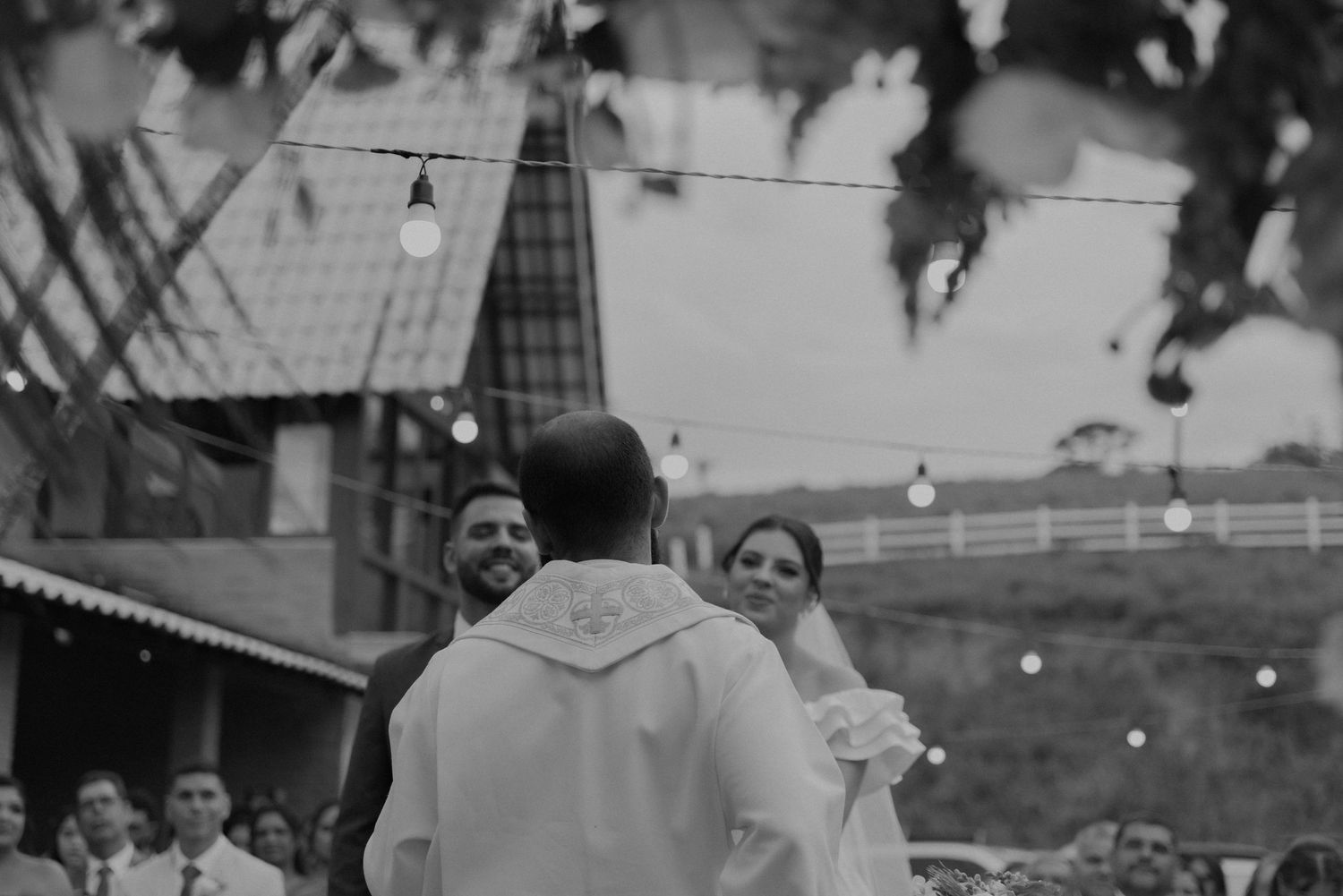 Black and white photo of newlyweds celebrating at an outdoor wedding reception with string lights overhead.