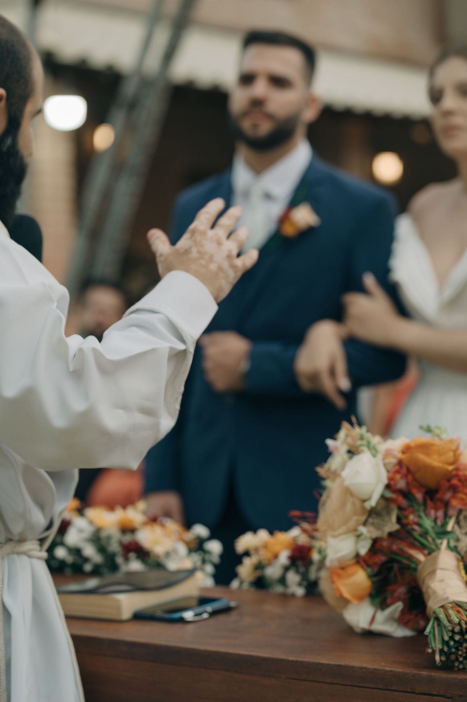 Priest gestures during wedding ceremony with floral arrangements on wooden table.