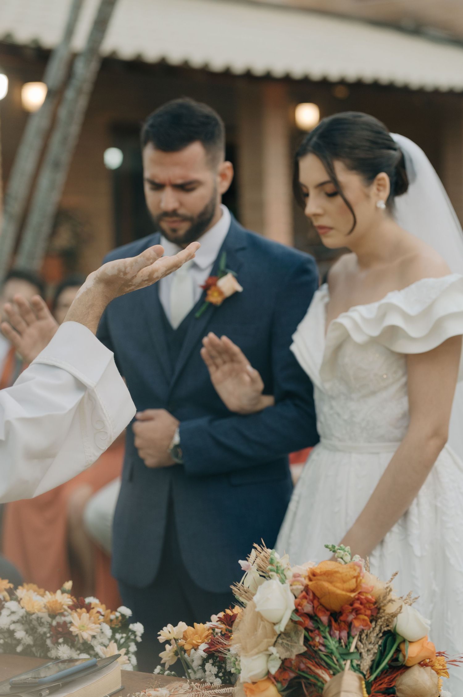 A priest blesses a couple during their wedding ceremony surrounded by floral arrangements.