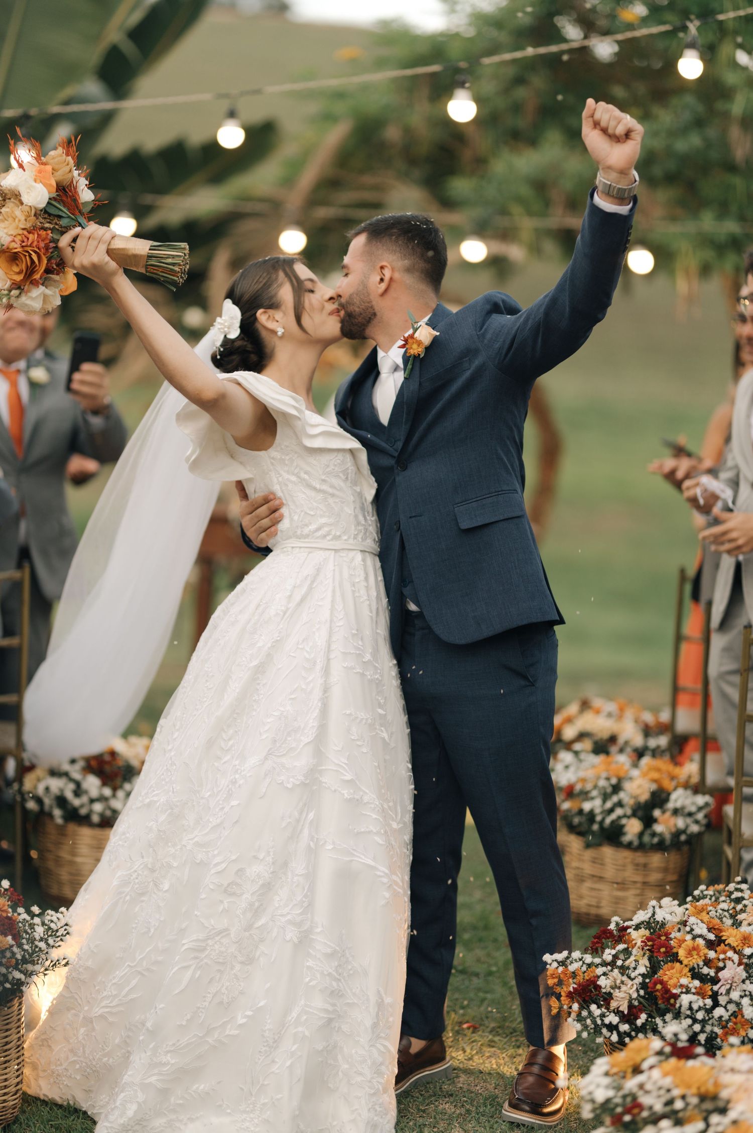 Triumphant wedding kiss with raised arms as guests celebrate under string lights.