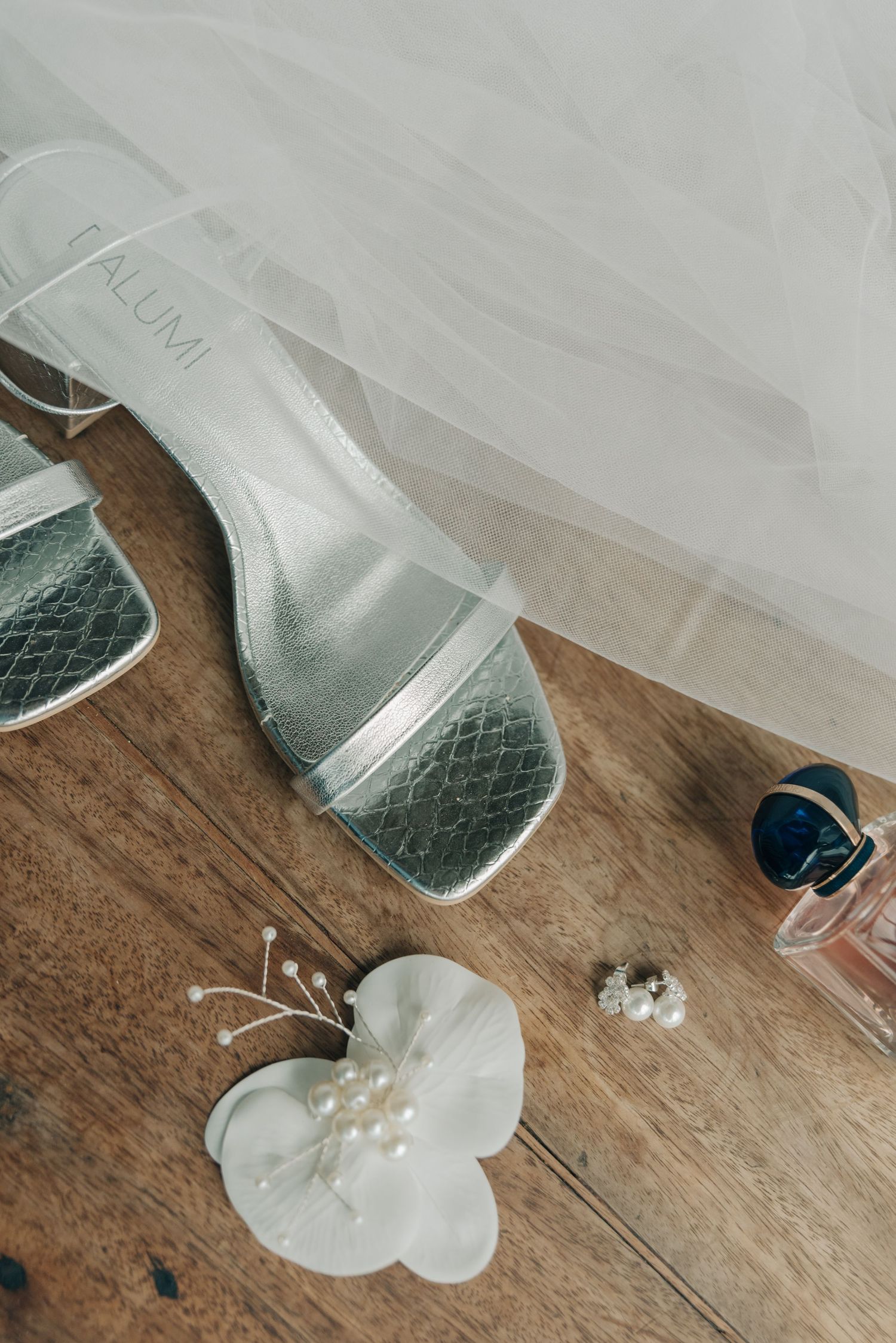 Wedding shoes and white floral accessories on wooden surface with delicate veil.