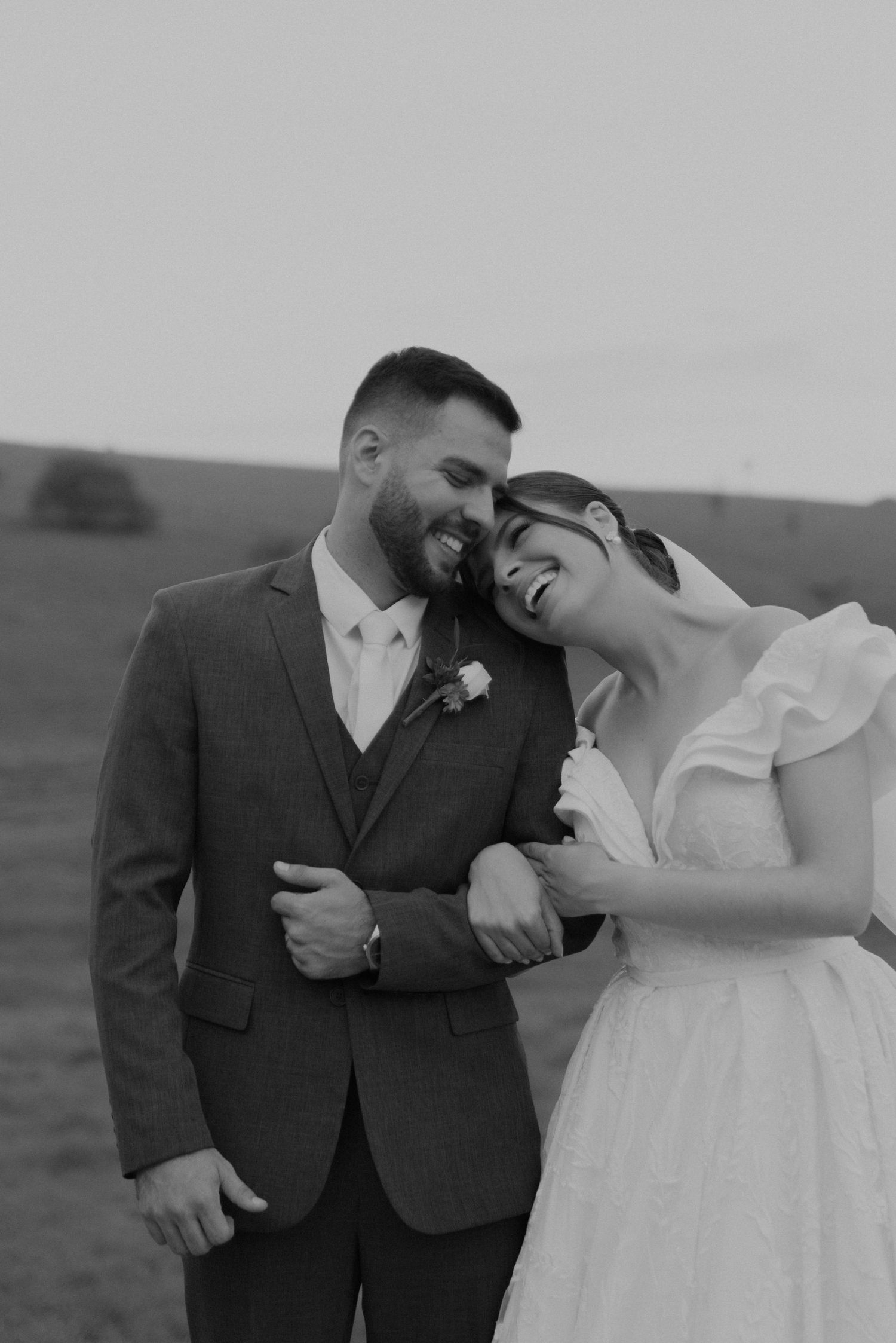 Black and white portrait of a wedding couple laughing together against a rural landscape.