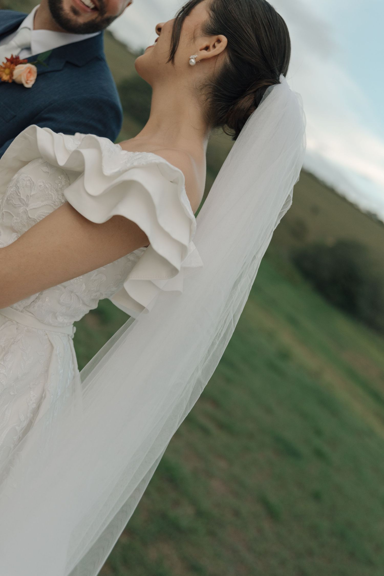 Close-up detail of a bride's ruffled wedding dress and flowing veil against a pastoral backdrop.