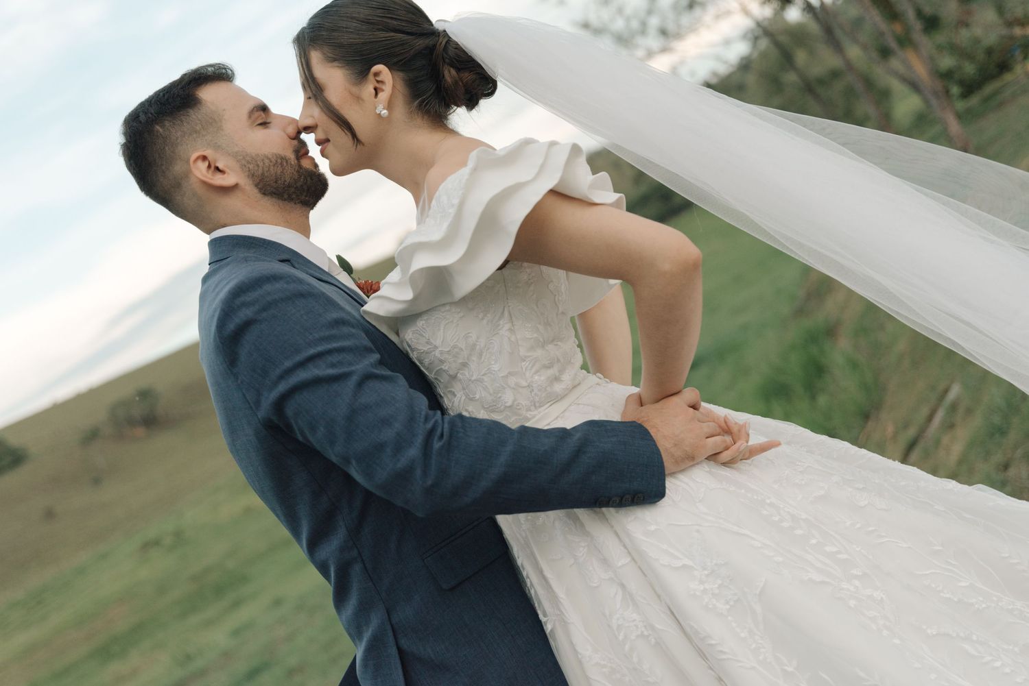 Romantic wedding portrait of couple embracing with bride's veil flowing in outdoor setting.