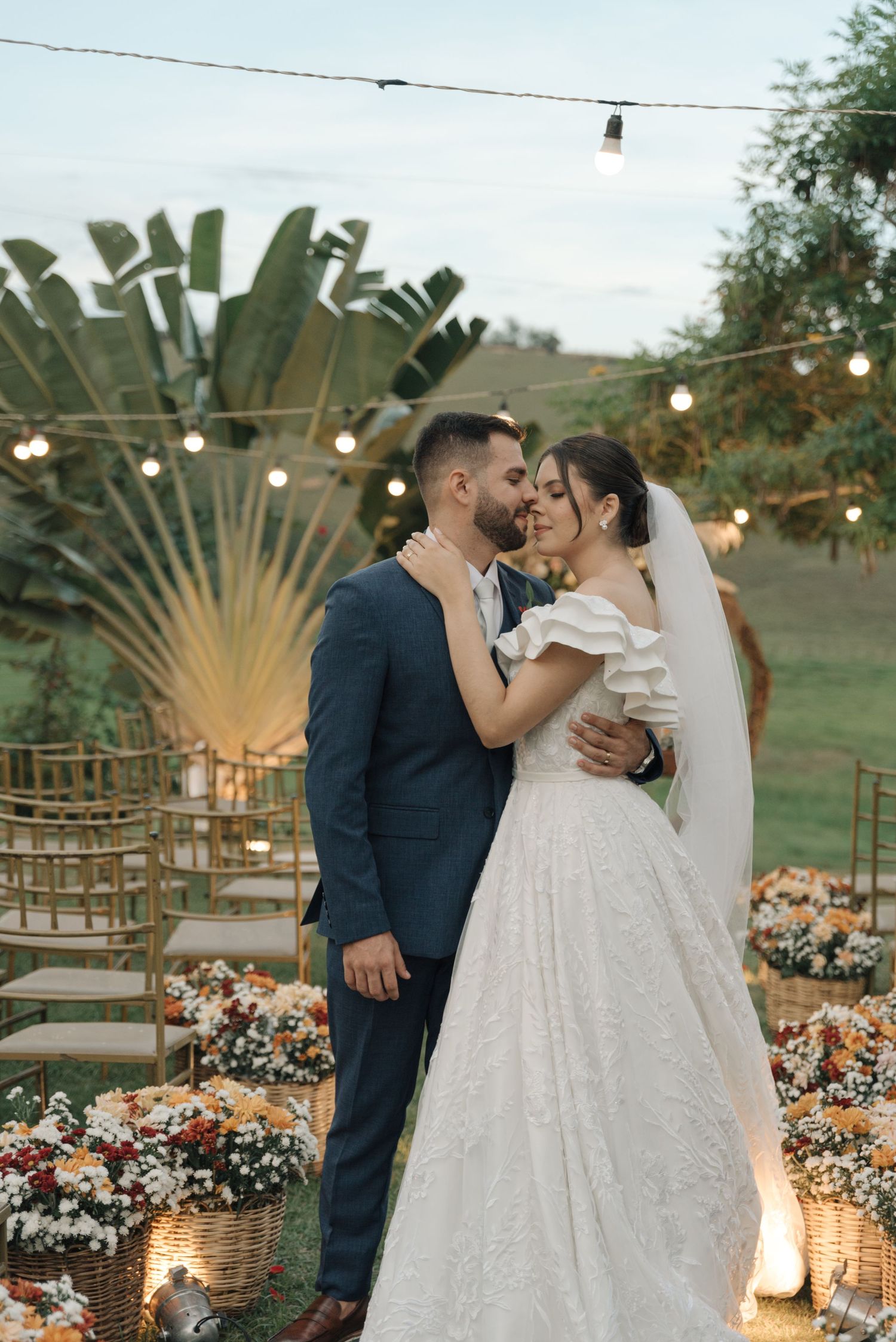 A newlywed couple shares a romantic moment in a garden setting with string lights and tropical foliage.