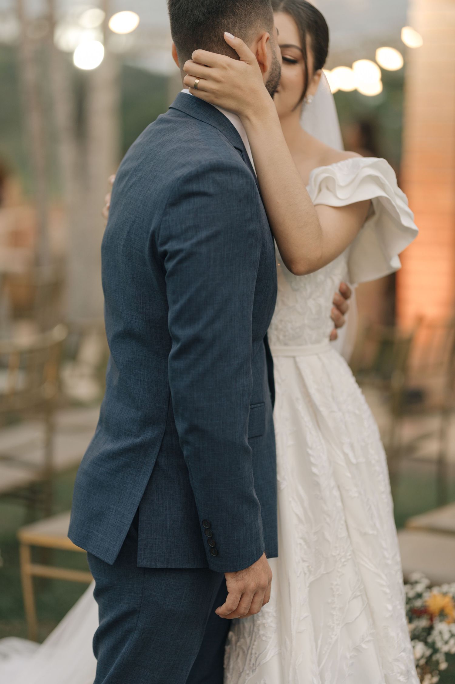 A romantic wedding embrace captured in soft evening light with string lights in the background.