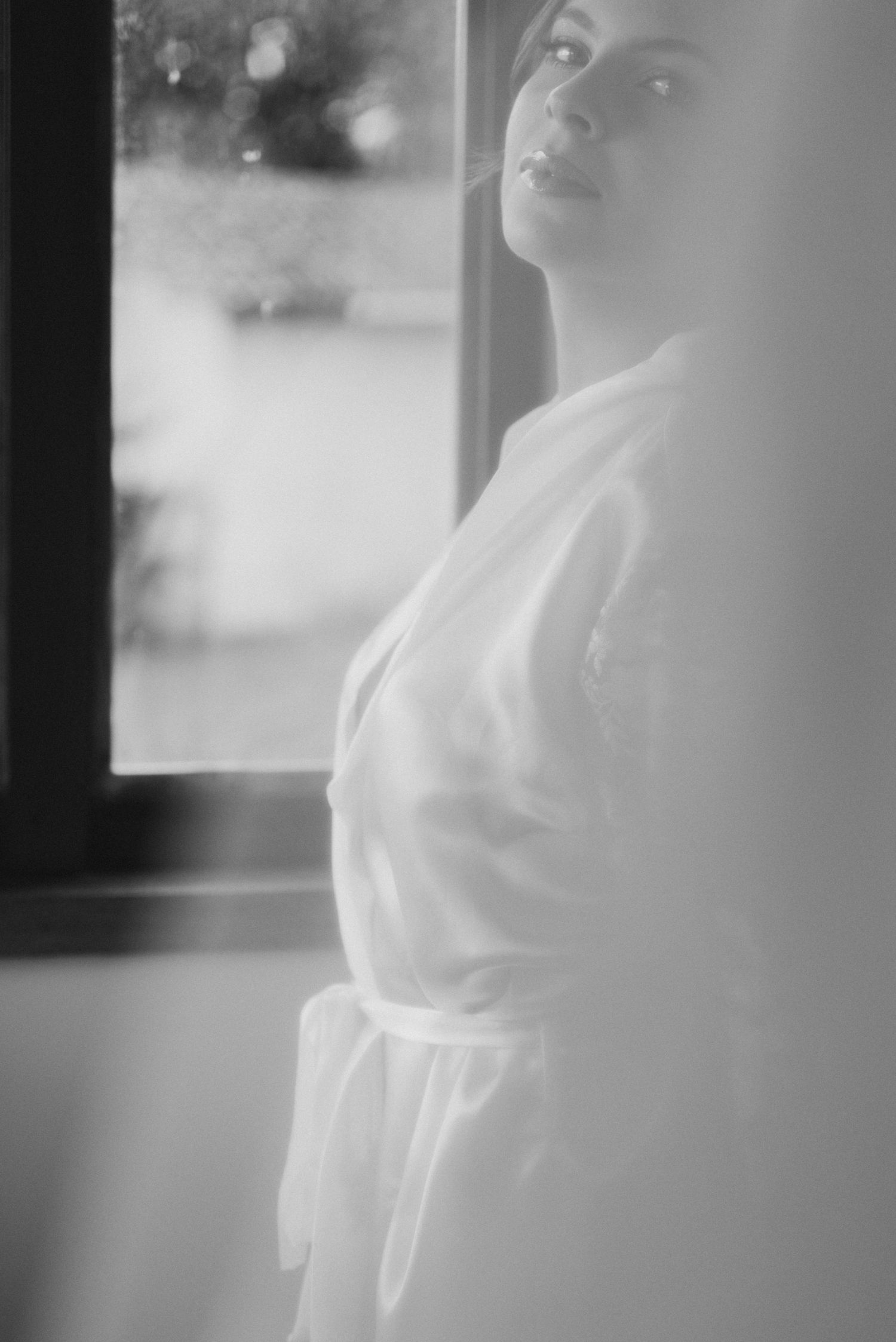 Dreamy monochrome portrait by window showing bridal preparation in white robe.