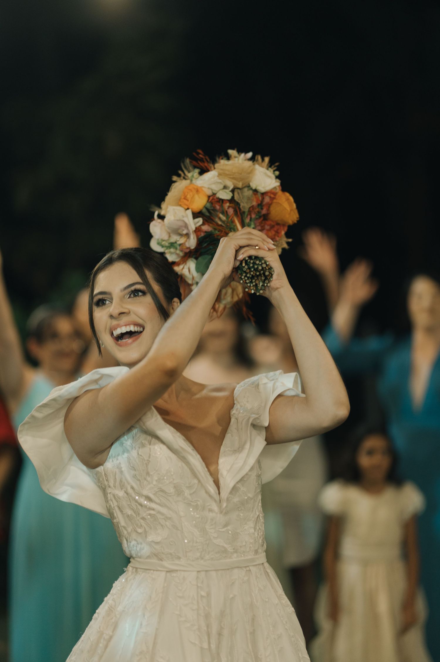 A bride in white dress joyfully holds up her wedding bouquet during the reception celebration.