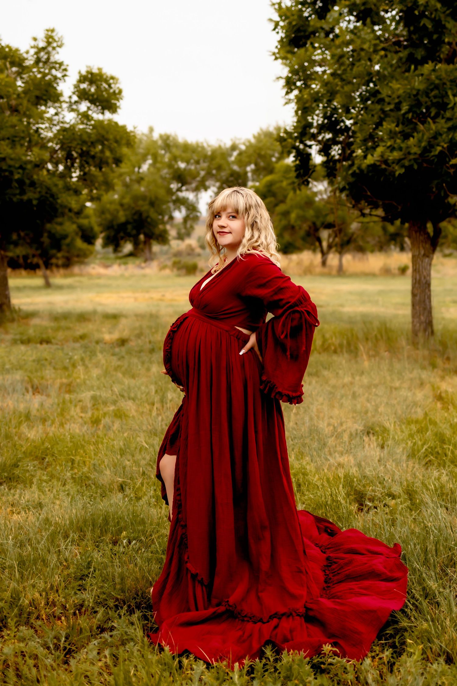 A maternity portrait features a woman in a deep red velvet gown standing in a rural setting.