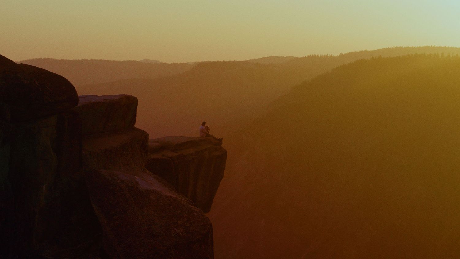 Silhouette of person on cliff edge against golden mountain landscape at sunset.