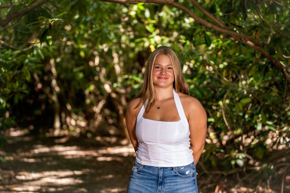 Person in white tank top and jeans standing against green foliage background.