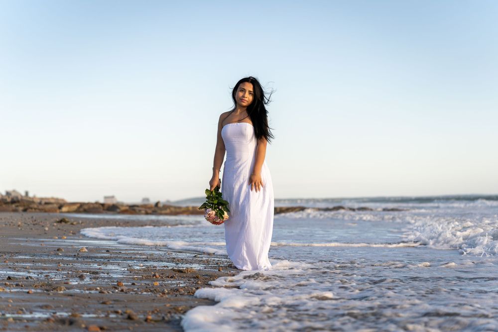 Person in flowing white dress standing at ocean's edge during golden hour with waves lapping at their feet.