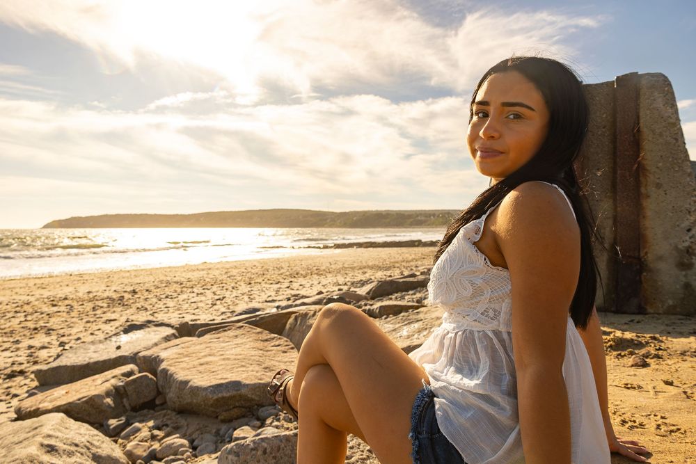 Person in white dress sitting on coastal rocks during sunset at beach.
