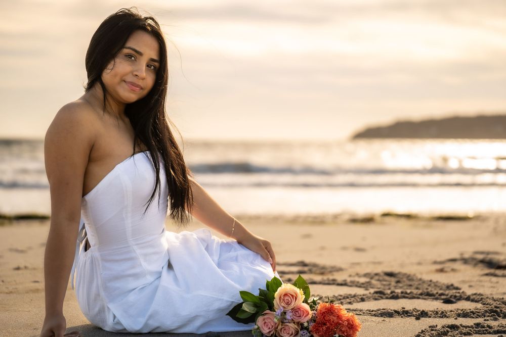 Person in white dress sitting on beach at sunset with bouquet of flowers.