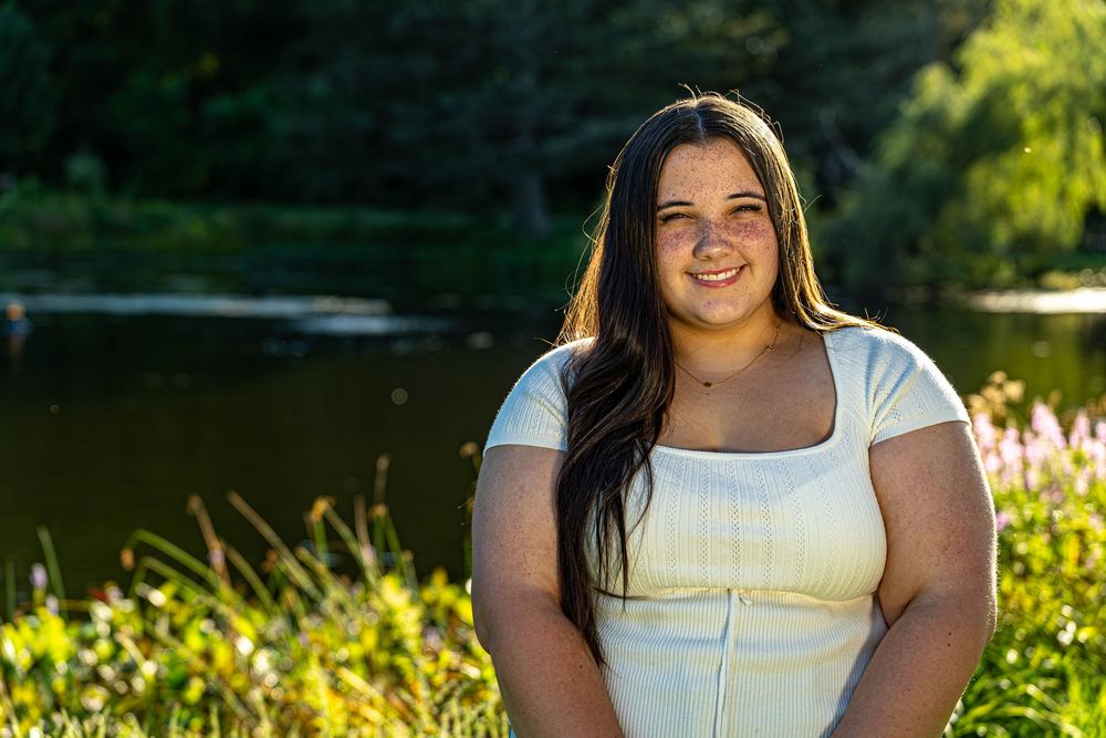 Person in yellow top poses outdoors with lake and greenery in background.