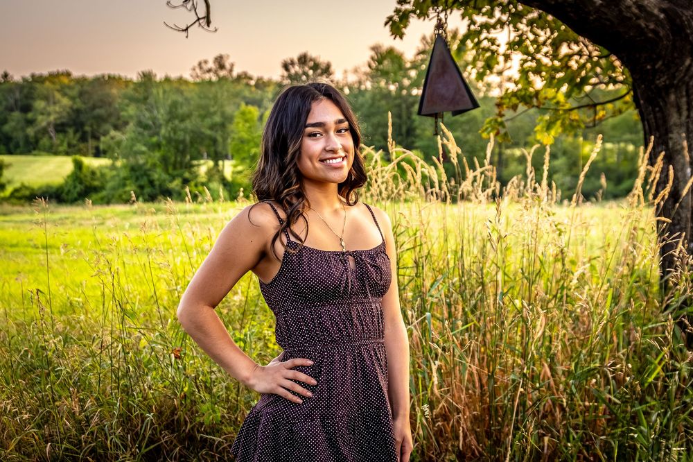 Person in a brown dress poses gracefully in a grassy field during sunset.